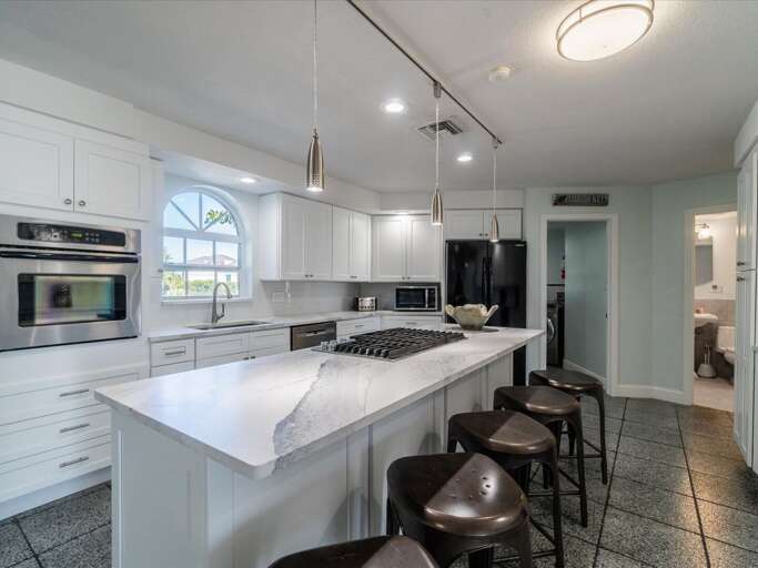 Brightly Lit Kitchen With White Counters And Stools, Leading Into Living Space